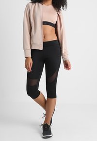 Nainen, joka on pukeutunut vaaleanpunaiseen vetoketjulliseen takkiin, samansävyiseen urheiluliiviin, mustiin verkkopaneelisiiroihin legginsseihin ja mustiin urheilukenkiin, seisoo jalat ristissä.