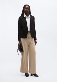 Must ülikonnajakk valge kraega särgi peal, ühendatud beežide laiade pükstega ja mustade kontsakingadega. Hoides käes väikest musta kotti.