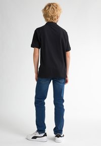 Svart pikétröja med korta ärmar, i kombination med blå jeans och vita sneakers med svarta detaljer, sedd bakifrån.