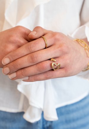 Zwei sanft ineinander verschränkte Hände, die goldene Ringe und ein goldenes Armband tragen, mit einer weißen Bluse und blauen Jeans im Hintergrund.