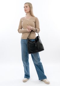 Vrouw met blond haar die een beige geribbelde cardigan en een blauwe spijkerbroek draagt, en met één hand een grote zwarte leren handtas vasthoudt.