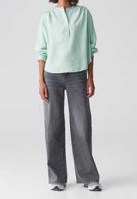 Blusa verde menta, con texture, colletto a metà bottoni e maniche arricciate, abbinata a jeans grigi sbiaditi a gamba larga e sneakers.