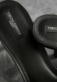 Sandales en cuir noir avec une texture lisse et un bout arrondi. Présente une étiquette "TABITHA SIMMONS NEXT" sur la semelle intérieure. Design minimaliste.