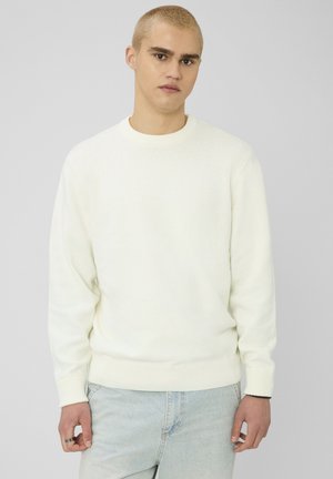 Jeune homme aux cheveux blonds courts portant un pull en maille blanc et un jean bleu clair, debout devant un fond uni gris clair.