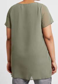 Olijfgroene, doorschijnende blouse met korte mouwen, ronde halslijn, langere achterkant en een gladde textuur. G gedragen over geruite broek.