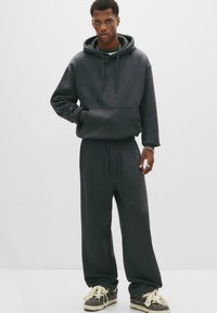 Ung man i en mörkgrå oversized huvtröja, matchande vidbyxor och sneakers, stående mot en enkel vit bakgrund.