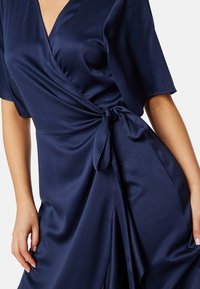 Navy blauwe wikkeljurk van gladde satijn, met korte mouwen, een strik bij de taille en een vloeiende silhouette met gedrapeerde stof.