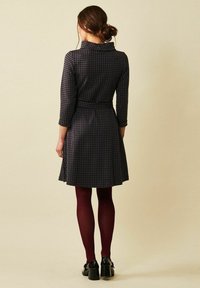 Musta-valkoinen houndstooth-kuviollinen mekko, jossa on kolmen neljänneksen hihat, vyötäröllä vyö, liehuva hame, yhdistettynä viininpunaisiin sukkahousuihin ja mustiin kenkiin.
