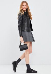 Zwarte leren motorjack met ritsdetails, grijze geruite rok, zwarte lakleren loafers en een kleine zwarte handtas met een gedraaid handvat.