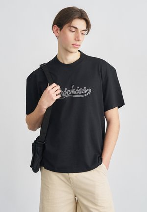 Mladý muž v čiernom tričku značky "Dickies", béžových nohaviciach a s čiernou crossbody taškou, pózuje na jednoduchom svetlošedom pozadí.