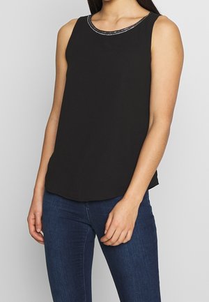 Kvinde iført sort ærmeløs top med perlebesat halsudskæring og mørkeblå denimjeans, stående mod en ensfarvet lys baggrund.