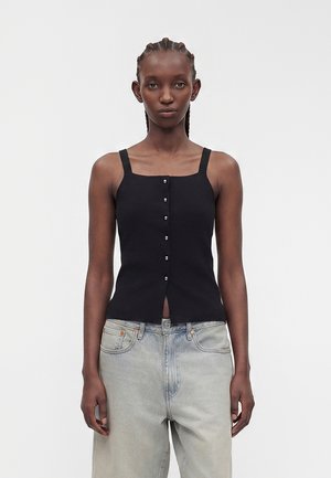 Kvinde med fletninger har sort ærmeløs knap-top og lyse afslappede jeans mod en ensartet lys baggrund.