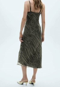 Robe maxi en tissu transparent, de couleur vert olive avec un motif ondulé noir. Dotée de fines bretelles et d'une coupe fluide. Jambes visibles, associée à des talons crème.