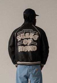 Black leather jacket with embroidered beige text "STATE OF MIND" on the back. Features ribbed cuffs and hem, contrasting white stitching.