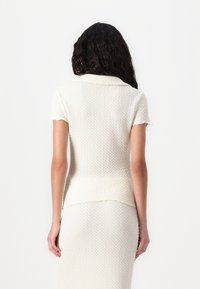 Top tricotat cu mânecă scurtă, de culoare crem, care prezintă un model texturat, guler pliat și o siluetă ușor evazată la tiv.