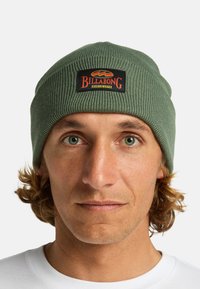 Berretto a coste verde con una patch rettangolare nera che presenta il logo Billabong, indossato da una persona con capelli ricci di lunghezza media.