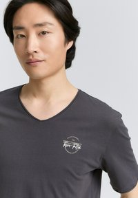 Mörkgrå t-shirt med v-ringning och korta ärmar, med en rund logotyp och textdesign i vitt på vänster bröstområde. Slät tygtextur.