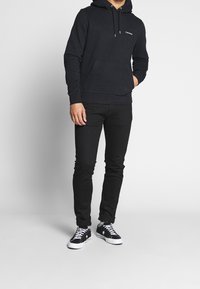 Felpa nera con tasca a canguro, coulisse regolabile, dettaglio del logo sul petto. Jeans neri slim-fit, abbinati a sneaker nere.