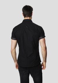 Camisa negra de manga corta con cuello con solapa, que presenta una textura suave y puños con patrón en contraste. Llevada con jeans oscuros, vista desde atrás.