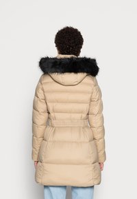 Béžový prošívaný kabát s páskem v pase, texturovaným prošíváním a černým lemováním kapuce z umělé kožešiny. Má dlouhé rukávy a střih do poloviny délky.