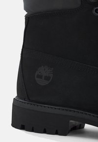 Botín negro impermeable hecho de gamuza con suela de goma. Presenta una superficie texturizada, cordones negros y un logo grabado en el lateral.