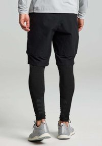 Pantaloni sportivi neri abbinati a leggins compressivi neri. Accenti testurizzati notevoli sui leggins e scarpe sportive grigie con motivo.