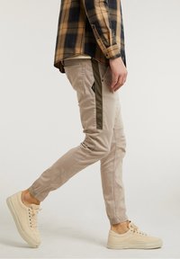 Beige slim-fit byxor med olivgröna sidosträck. Tillverkade i lättviktsmaterial, med dragkedjefickor och elastiska muddar. Matchat med beige sneakers.