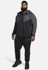 Schwarzer Nike-Trainingsanzug mit einer zip-up Kapuzenjacke im Farbblockdesign und rotem Logo, kombiniert mit passenden Jogginghosen und schwarzen Sneakers.