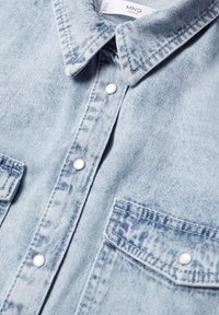 Chemise en denim bleu clair avec un col classique, deux poches poitrine et des boutons-pression blancs. Le tissu présente une finition douce et texturée.