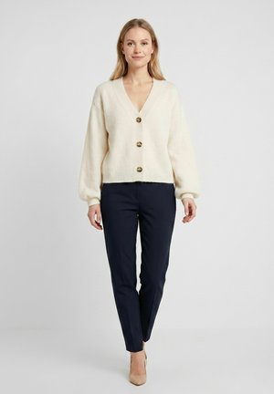 Femeie în picioare, purtând un cardigan bej cu nasturi, pantaloni drepți bleumarin și pantofi cu toc nude, pe un fundal simplu și deschis la culoare.