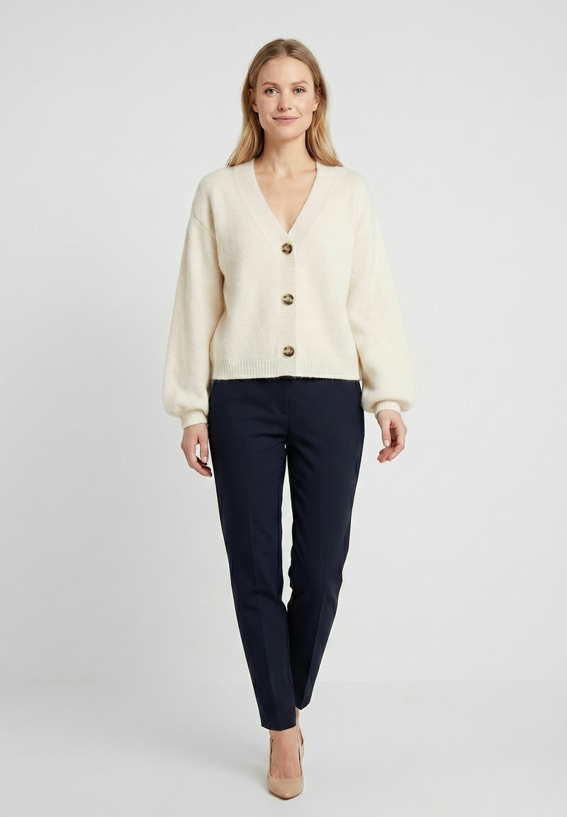Femme debout portant un cardigan beige boutonné, un pantalon droit bleu marine et des talons couleur nude sur un fond clair uni.