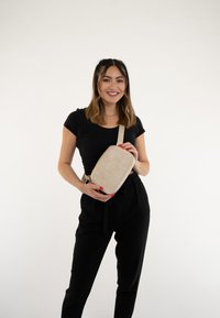 Beige Umhängetasche mit glatter Oberfläche und rechteckiger Form, ausgestattet mit einem Tragegurt, auf dem das Wort "TRIE" gedruckt ist. Das Modell hält die Tasche vor einem neutralen Hintergrund.