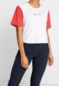 Osoba v bílém crop T-shiru s červenými rukávy a námořnickými úzkými kalhotami, stojící na jednobarevném pozadí.