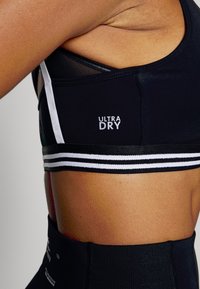 Top desportivo preto com riscas brancas e logótipo "ULTRA DRY". Apresenta um ajuste justo, textura suave e detalhes em malha para respirabilidade.