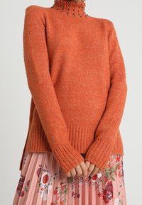 Maglione arancione lavorato a maglia con colletto alto decorato con perline scure. Presenta polsini e orlo a costine, abbinato a una gonna floreale plissettata.