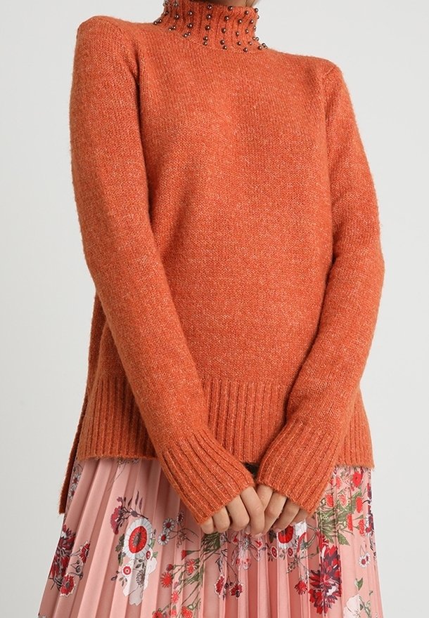 Maglione arancione lavorato a maglia con colletto alto decorato con perline scure. Presenta polsini e orlo a costine, abbinato a una gonna floreale plissettata.