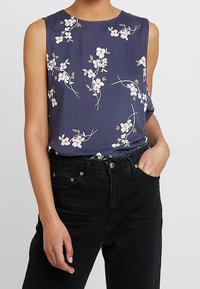 Marinblå ärmlös topp med vita blommönster och gröna blad. Gjord av lättviktig tyg, i kombination med svarta högmidjade jeans.