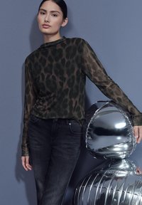 Doorzichtig groen topje met luipaardprint en een hoge hals, gecombineerd met donkere spijkerbroeken. Er staat een reflecterend zilveren beeldje in de buurt.