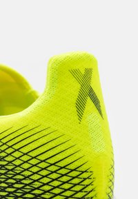 Kenkä, jossa on neonkeltaiset urheilukengät, joissa on teksturoitu verkko, mustat diagonaaliset raidat ja "X"-logo. Tarjoaa tyylikkään, modernin ilmeen.