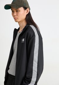 Jaqueta preta com fecho zip e detalhes em branco nas mangas, com o logo da Champion. Usada sobre uma camisa cinza, complementada por um boné verde.