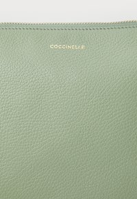 Green leather pouch with a textured finish, featuring a gold embossed brand name "Coccinelle" at the top and an exposed zipper closure.
