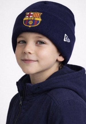 Młody chłopiec w granatowej czapce FC Barcelona i pasującej kurtce, lekko się uśmiechający, zwrócony przodem na jednolitym tle.