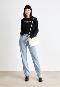 Sudadera negra con el logo "Calvin Klein Jeans", vaqueros rectos de tiro claro, bolso de hombro blanco y botas negras hasta el tobillo. Fondo neutro.