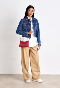 Borsa a spalla rossa, giacca di jeans, camicia bianca, pantaloni beige a gamba larga, sneaker chiare. Design semplice con texture lisce e minimi accenti.