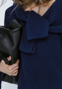 Marineblauer gestrickter Pullover mit einer großen Schleifen-Applikation am Ausschnitt; kombiniert mit einer glatten schwarzen Leder-Clutch, die Textur und Design betont.