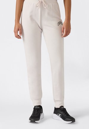 Persona che indossa pantaloni da jogging beige chiaro con coulisse in vita e sneakers bianco e nero, in piedi davanti a uno sfondo semplice.