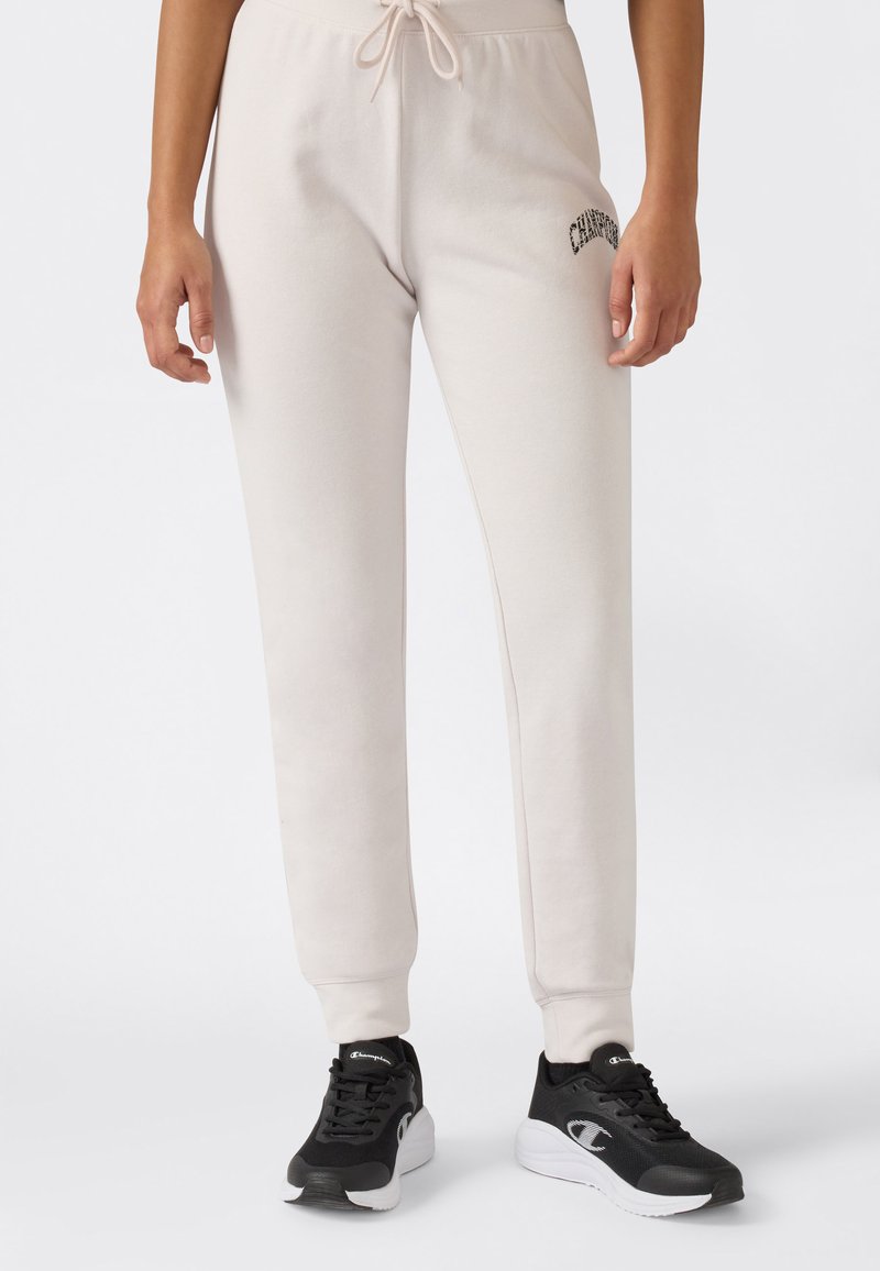 Persona che indossa pantaloni da jogging beige chiaro con coulisse in vita e sneakers bianco e nero, in piedi davanti a uno sfondo semplice.