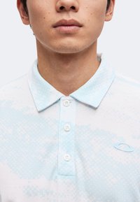 Jeune personne portant un polo bleu clair et blanc à motif, avec un col et trois boutons, montrant le bas du visage et le cou.