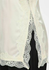 Tissu satin ivoire avec une bordure en dentelle florale délicate et des bords festonnés sur fond noir, montrant des motifs et la texture détaillés de la dentelle.