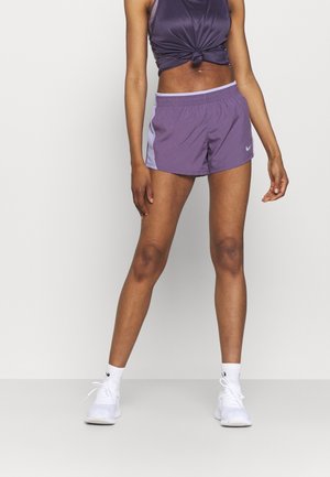 Kvinde iført lilla sportsshorts, lilla ærmeløs top bundet ved taljen, hvide sokker og hvide løbesko, står mod en ensfarvet baggrund.